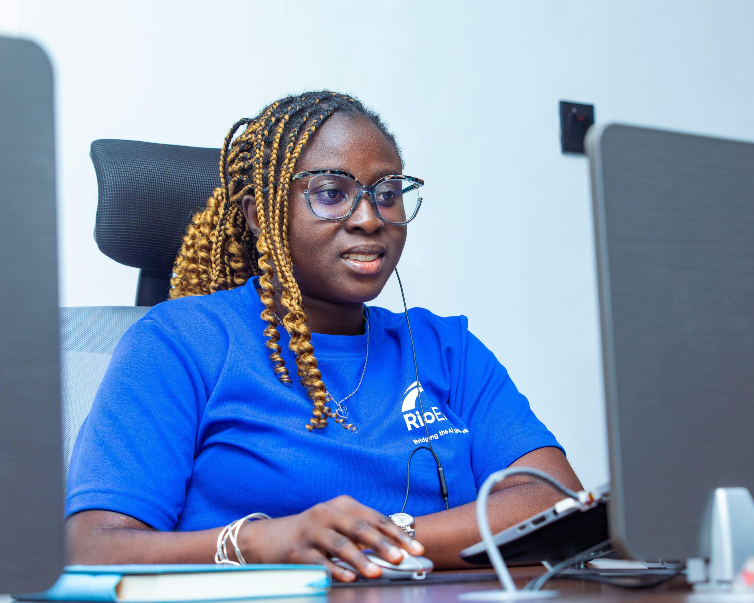 Confident woman in blue t-shirt working at a desk with multiple screens, illustrating modern office environment.